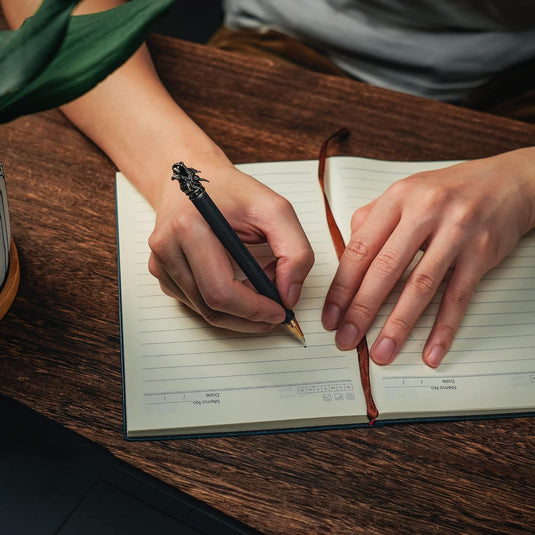 Persoon die in een notitieboekje op een houten tafel schrijft met een bril ernaast en een stijlvol en duurzaam schrijfapparaat.