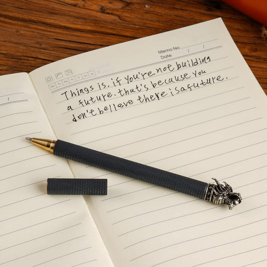 Zin met productnaam: Een minimalistisch design schrijfapparaat ligt op een open notitieboek met een handgeschreven citaat: "Het is zo dat je geen toekomst bouwt, dat komt omdat je niet gelooft dat er een toekomst is".